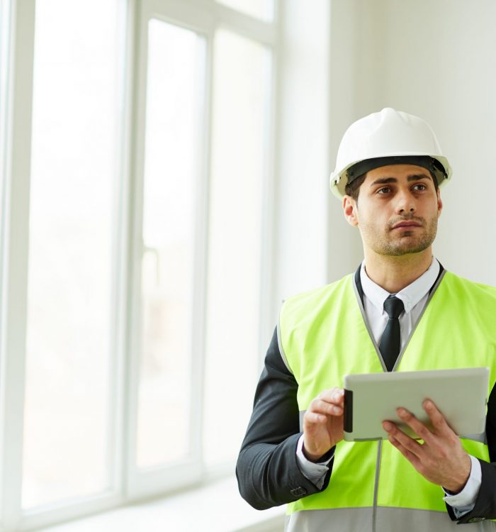 Waist up portrait of Middle-Eastern engineer wearing hardhat posing on construction site holding tablet, copy space